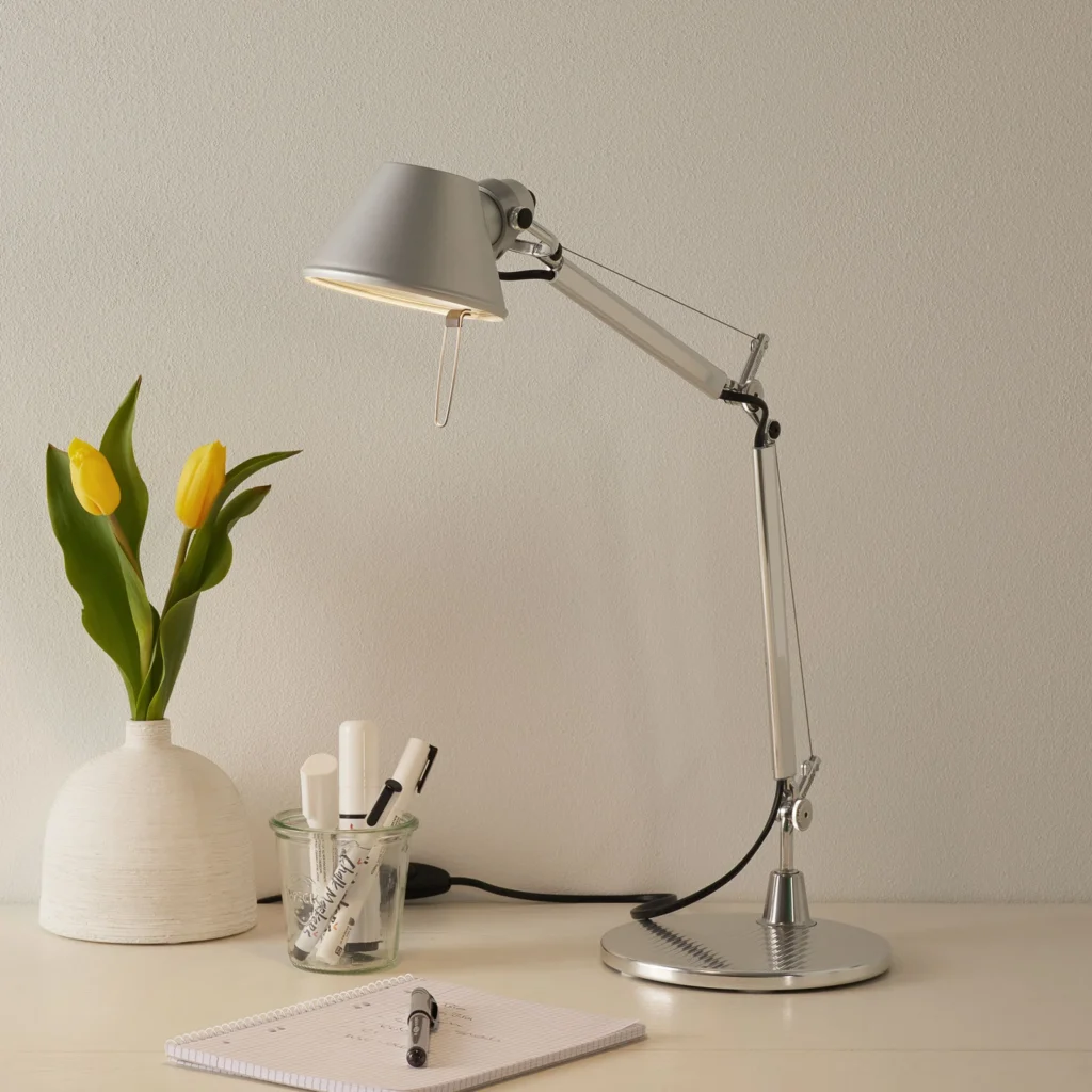 Silberne Artemide Tolomeo Micro Schreibtischlampe aus Aluminium auf einem hellen Tisch neben einer Vase mit gelben Tulpen und Schreibutensilien.